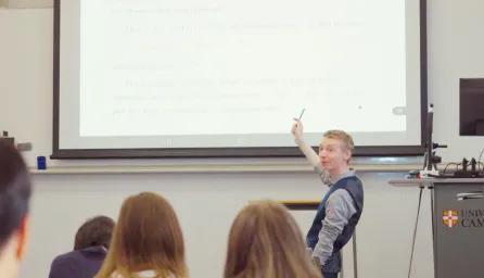 Academic stood at front of classroom pointing at projection of teaching materials