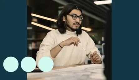 Man with shoulder length dark hair, sat at desk with pencil in hand