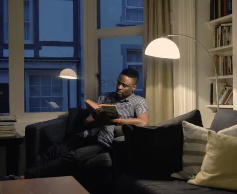 Man reading on a sofa in a dimly lit apartment, illuminated by a single overhead light