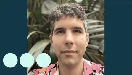 Man with greying dark curly hair, stood in front of large plant 