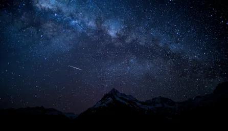 A starry night sky above dark mountain silhouettes, with one shooting star visible against the clear sky