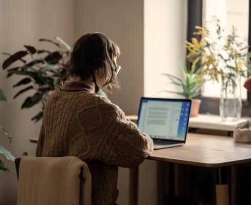 A woman wearing a brown jumper, working on a laptop at a wooden desk in a room with houseplants and natural light from a nearby window