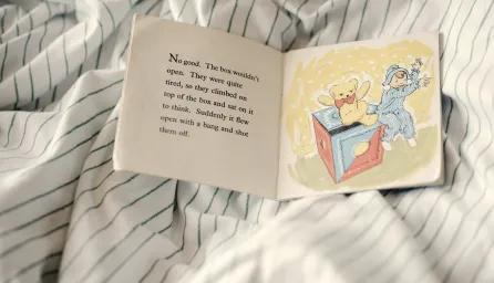 An open vintage children’s book lying on striped bed sheets, showing a scene from a British story with a teddy bear and child being launched from a toy box