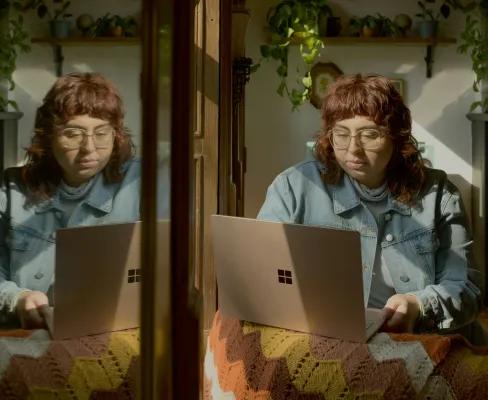 A person typing on a laptop at a table with a blanket, her reflection visible in the window opposite, with greenery and decorative plants behind them