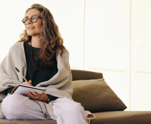 A woman holding a pen poised over a notepad, sitting cross-legged on a sofa in a softly lit room