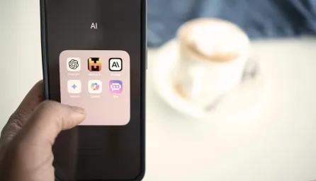 A person holding a mobile phone displaying AI-related apps including ChatGPT, with a blurred background showing a coffee on a table