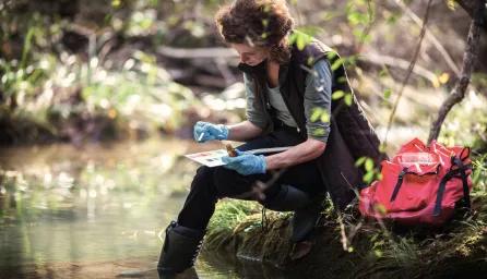 A woman testing lake water with a colour sample board, sitting on the grass with her feet in the lake, a red bag beside her and trees in the background