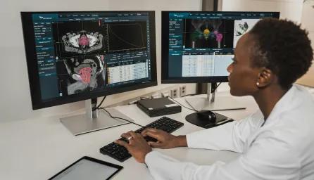 A researcher in a white coat analysing brain and body scans on dual monitors in a clinical setting