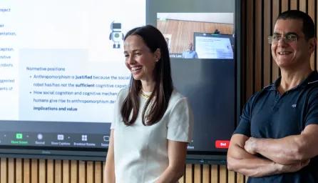 A man and a woman smiling and standing in front of a large wall-mounted display screen with a presentation on display.
