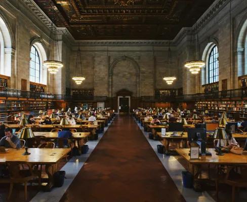 People studying at desks in a long corridor of a library, surrounded by bookshelves