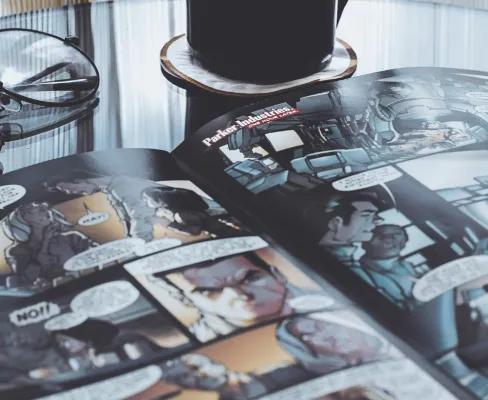 An open comic book lying on a glass table, with a black mug of coffee beside it and a pair of round glasses resting on the comic’s pages.