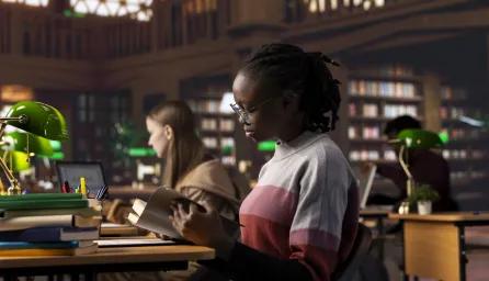 Students reading and working at desks in a quiet library setting, surrounded by shelves of books