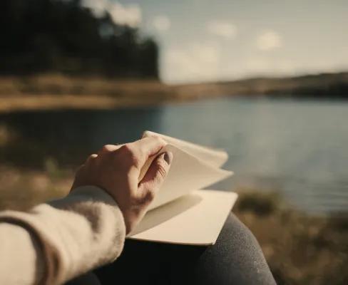 A person’s hand turning a page in a notebook while sitting by a lake, with blurred trees and sky in the background