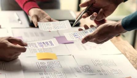 Several hands pointing to a mind map covered in post-it notes during a brainstorming session, with more notes and leaflets being held