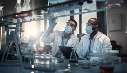 Two scientists wearing masks, lab goggles, and white coats examining a cell culture plate in a laboratory, conical flasks and test tubes are blurred in the background.