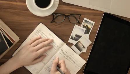 A flat lay image showing a person's hands writing in a planner or journal open to "FEBRUARY" on a wooden desk. To the left are stacked books, including one titled "YOUR SOUL" by Nikita Gill. To the right are a tablet, a pair of eyeglasses, a white coffee cup, and several small instant photo prints scattered on the desk.