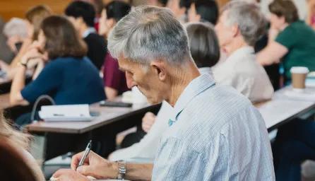 Mature male student taking notes in a lecture theatre surrounded by other students.