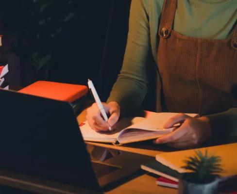 A person is writing in a notebook at a cluttered desk with a laptop and many books, under the light of a desk lamp.