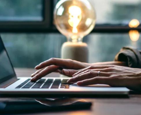 Side view of an open laptop on a table and a person working on it, a glowing lamp in the background.
