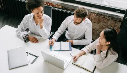 Three colleagues collaborate at a office table, focused on a laptop and notebooks.