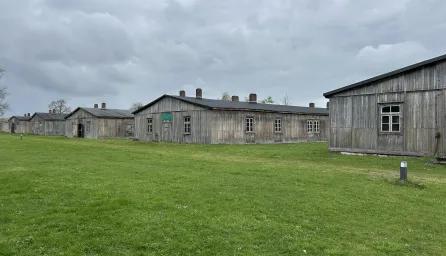 An image featuring several weathered wooden barracks arranged in rows on a green lawn, under an overcast sky.