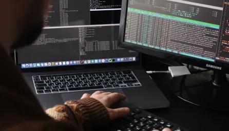 Close up from over the shoulder of several computer screens showing coding and someone working at a keyboard.