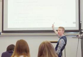 Academic stood at front of classroom pointing at projection of teaching materials