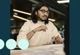 Man with shoulder length dark hair, sat at desk with pencil in hand