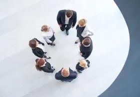 Group of 8 people pictured from above, dressed in business clothing and standing in a circle. 