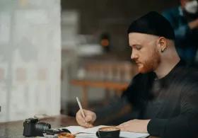 A man writing in a notebook at a table in a coffee shop, with a camera beside him and a blurred background