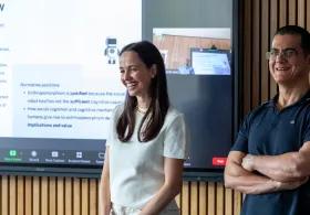 A man and a woman smiling and standing in front of a large wall-mounted display screen with a presentation on display.