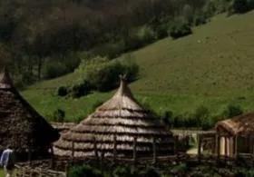A painting of rustic wooden huts with thatched roofs set against green hills, trees, and dense woodland in the background, with a lone man walking away into the scene.