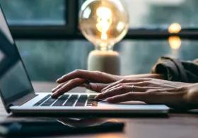 Side view of an open laptop on a table and a person working on it, a glowing lamp in the background.