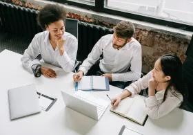 Three colleagues collaborate at a office table, focused on a laptop and notebooks.