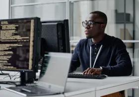 Young developer in eyeglasses concentrating on his online work on computer sitting at workplace