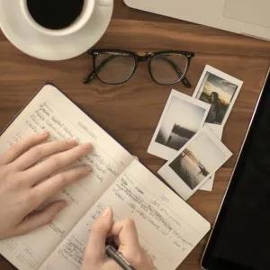 A flat lay image showing a person's hands writing in a planner or journal open to "FEBRUARY" on a wooden desk. To the left are stacked books, including one titled "YOUR SOUL" by Nikita Gill. To the right are a tablet, a pair of eyeglasses, a white coffee cup, and several small instant photo prints scattered on the desk.
