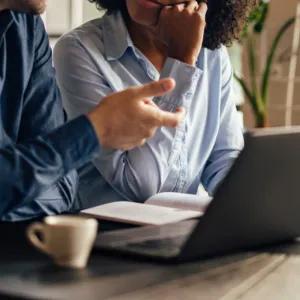 A man and a woman sitting together at an open laptop on a table, having a conversation.