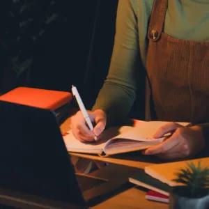A person is writing in a notebook at a cluttered desk with a laptop and many books, under the light of a desk lamp.