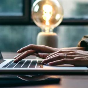 Side view of an open laptop on a table and a person working on it, a glowing lamp in the background.