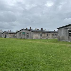 An image featuring several weathered wooden barracks arranged in rows on a green lawn, under an overcast sky.