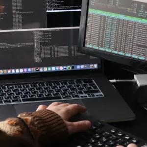 Close up from over the shoulder of several computer screens showing coding and someone working at a keyboard.