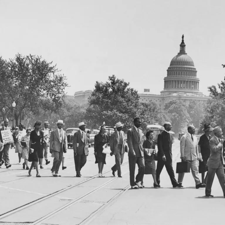 A black and white image of people marching in a protest, one holding an American flag, with trees and buildings in the background