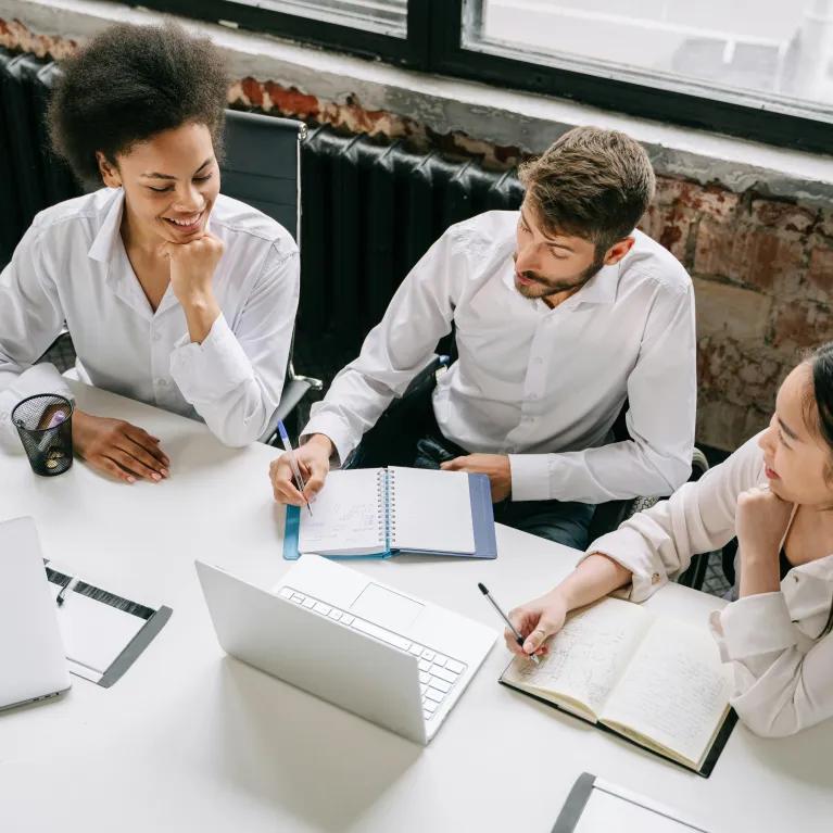 Three colleagues collaborate at a office table, focused on a laptop and notebooks.
