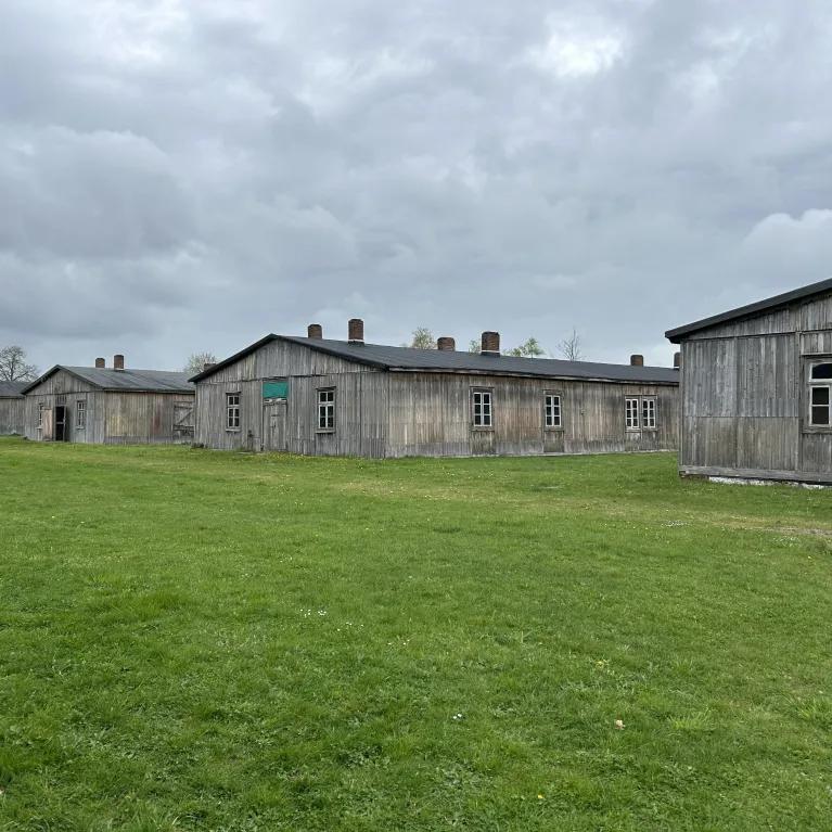 An image featuring several weathered wooden barracks arranged in rows on a green lawn, under an overcast sky.