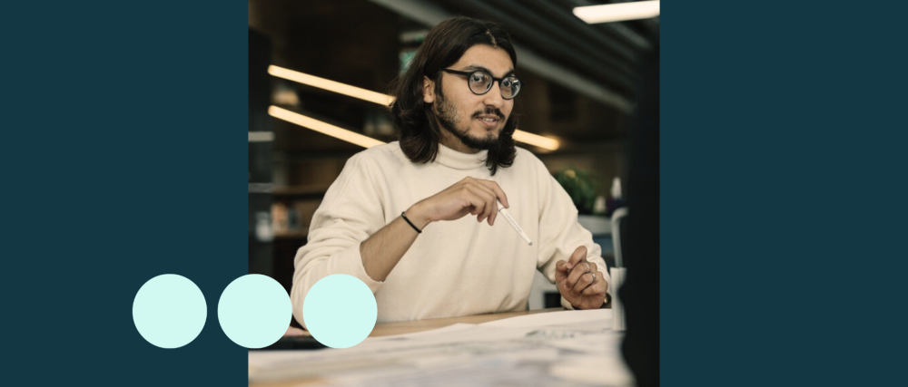 Man with shoulder length dark hair, sat at desk with pencil in hand