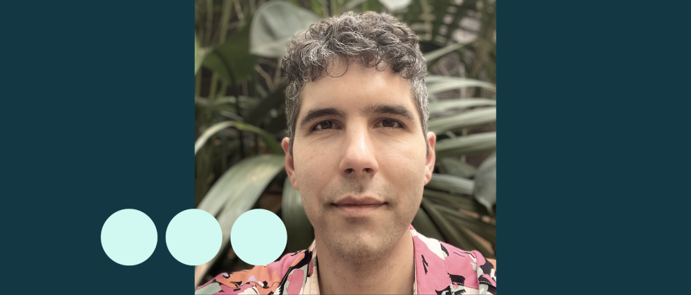 Man with greying dark curly hair, stood in front of large plant 